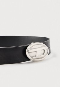 Cinto de couro preto com fivela oval prateada que apresenta um logótipo "TD" em relevo, exibido contra um fundo branco simples.