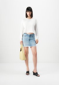 Kvinne iført en hvit langermet crop topp, lys blå slitt denimskjørt, svarte kilehæler, og holder en gul håndveske.