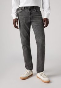 Jeans gri închis cu croi drept, având o textură subtilă și un design cu cinci buzunare, asociate cu adidași albi și o cămașă alba cu mânecă lungă.