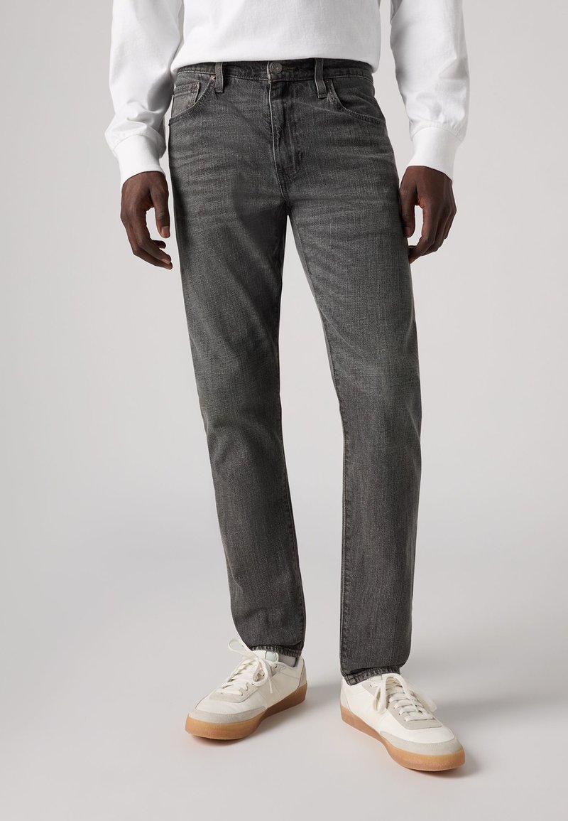 Jeans gris foncé à coupe droite, présentant une texture subtile et un design à cinq poches, accompagnés de baskets blanches et d'une chemise à manches longues blanche.