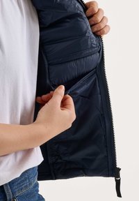 Une personne tient ouverte une veste matelassée bleu marine, montrant la poche intérieure avec sa main, portant un jean bleu et une chemise blanche.