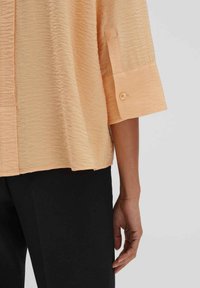 Blusa color pesca con una finitura texturizzata e stropicciata, maniche a tre quarti e un accento di bottone singolo sui polsini. Pantaloni neri visibili.