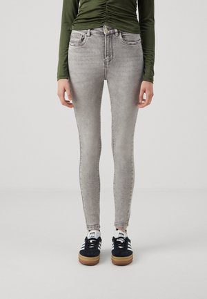 Person trägt hellgraue Skinny-Jeans, schwarz-weiße Sneakers und ein langärmeliges, olivgrünes Oberteil mit Raffung vor einem schlichten Hintergrund.