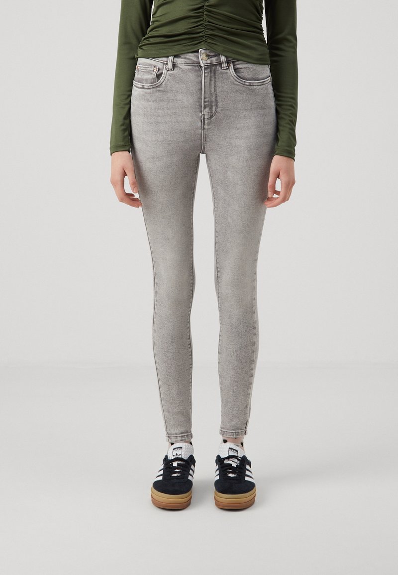 Personne portant un jean skinny gris clair, des baskets noires et blanches, et un haut à manches longues vert olive froncé sur un fond uni.
