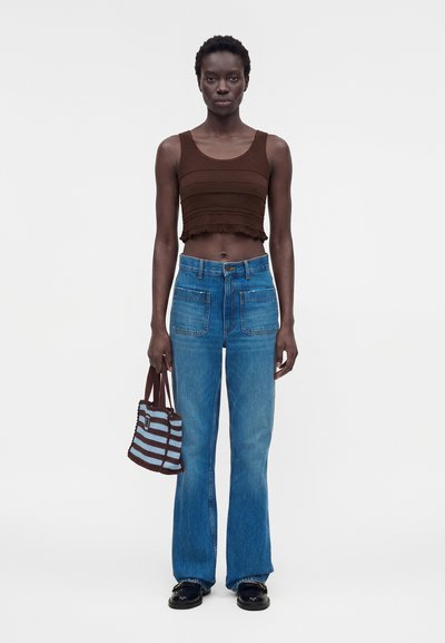 Γυναίκα με καφέ cropped τοπ, ψηλόμεσα μπλε τζιν, μαύρα παπούτσια, κρατώντας ριγέ καφέ και μπλε τσάντα, στέκεται μπροστά σε λευκό φόντο.