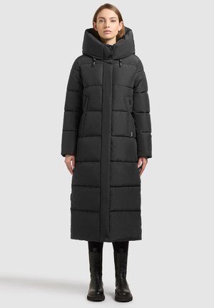 Donna che indossa un lungo cappotto invernale nero trapuntato con colletto alto, tasche frontali e stivali neri, in piedi davanti a uno sfondo semplice.