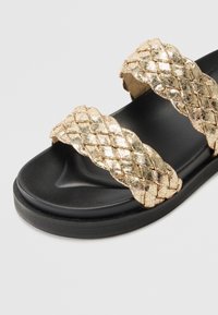 Sorte sandaler med svart farge som har to brede, metalliske gullvevde stropper med en kviltet tekstur, plassert på en formet svart såle.