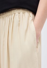 Άτομο που φοράει μπεζ παντελόνι με ελαστική μέση, με το ένα χέρι στην τσέπη και μαύρο κορμάκι ορατό στο ύψος της μέσης.