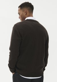 Brūns adīts džemperis ar brīvu piegriezumu, ribotām aprocēm un apakšmalu, ar V veida kakla dizainu un gludu tekstūru. Nēsāts virs baltas krekla.