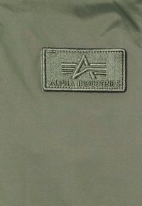 Vihreä kangas, jossa on ommeltu suorakulmainen läppä, jossa on teksti "ALPHA INDUSTRIES" ja logo, jossa on kulmikkaita viivoja ja raitoja.