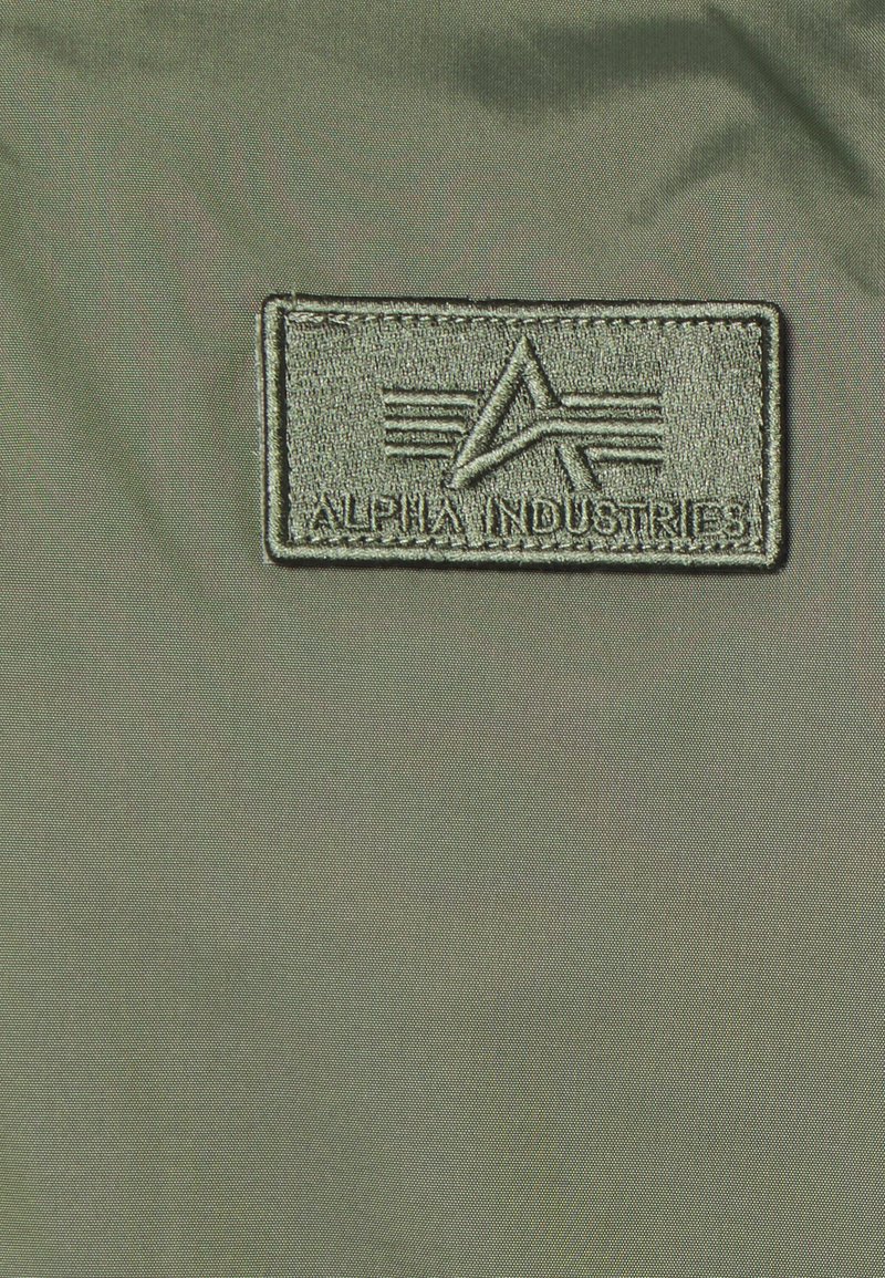 Vihreä kangas, jossa on ommeltu suorakulmainen läppä, jossa on teksti "ALPHA INDUSTRIES" ja logo, jossa on kulmikkaita viivoja ja raitoja.