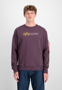 Ljubičasti dukserica s zlatnim logotipom "ALPHA INDUSTRIES" na prednjoj strani, rebrasti ovratnik i crvena oznaka detalj na rukavu.