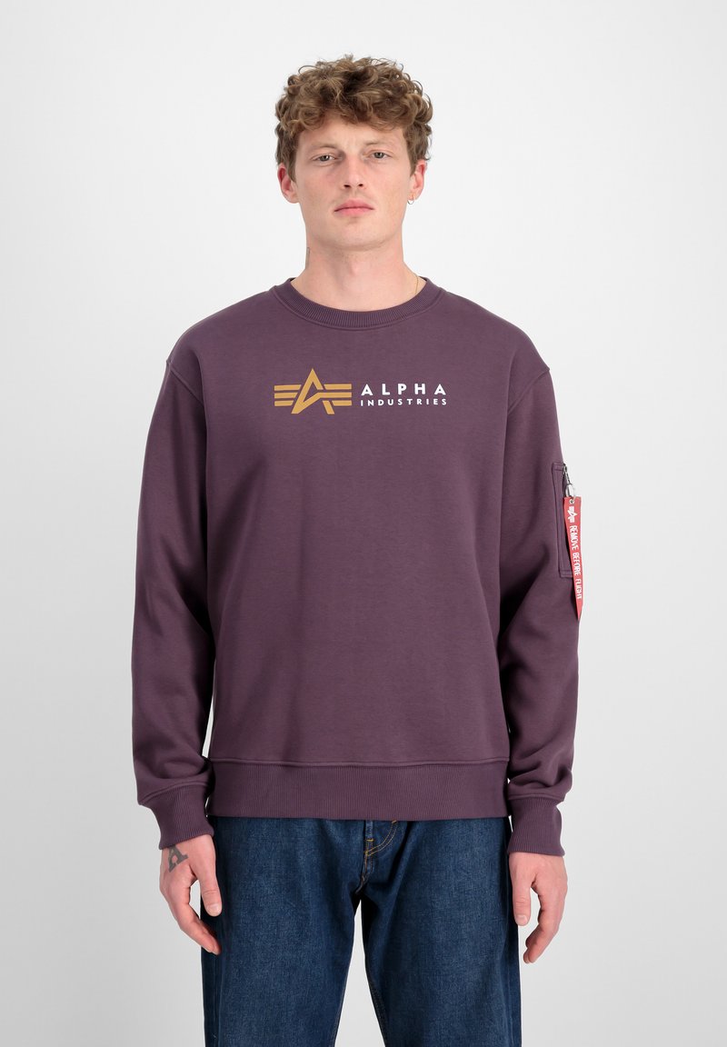 Ljubičasti dukserica s zlatnim logotipom "ALPHA INDUSTRIES" na prednjoj strani, rebrasti ovratnik i crvena oznaka detalj na rukavu.