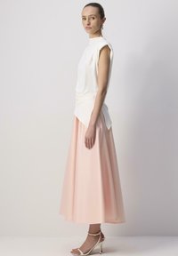 Haut sans manches blanc avec un design drapé associé à une longue jupe fluide rose pastel. La mannequin porte des talons transparents.