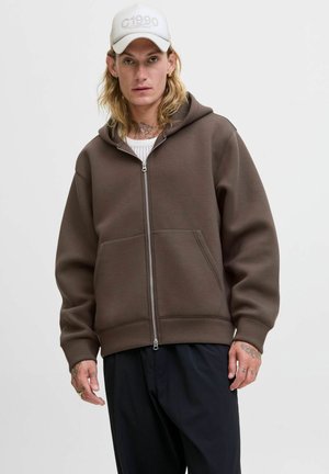 Sudadera marrón con cremallera y capucha grande, bolsillos frontales y puños acanalados. Combinada con pantalones negros y una gorra clara que tiene "C1990".