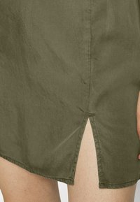 Saia verde-oliva com corte reto e fenda lateral. O tecido parece suave com um ligeiro brilho. A costura é visível ao longo da bainha.