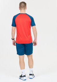 Man som står med ryggen mot kameran, iklädd en sportdräkt i röd och blå färg, vita strumpor och svartvita träningsskor, framför en vit bakgrund.