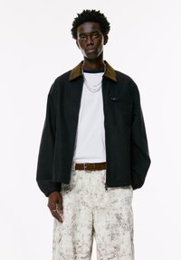 Black jacket with a brown corduroy collar, zip closure, and elastic cuffs, worn over a white T-shirt and light patterned cargo pants.
