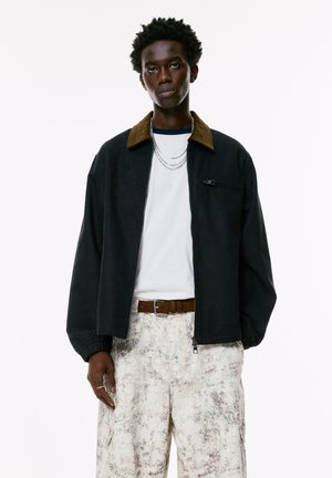 Black jacket with a brown corduroy collar, zip closure, and elastic cuffs, worn over a white T-shirt and light patterned cargo pants.