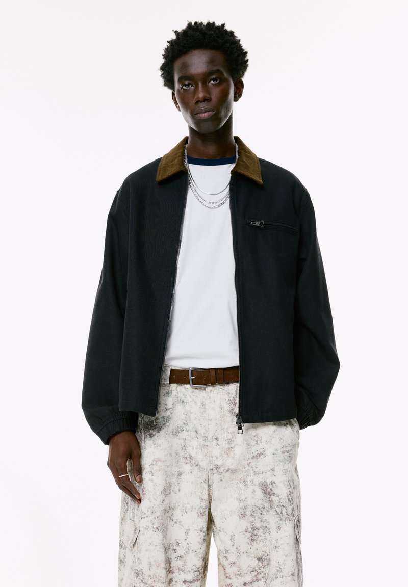 Black jacket with a brown corduroy collar, zip closure, and elastic cuffs, worn over a white T-shirt and light patterned cargo pants.