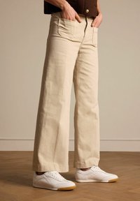 Beige wijde jeans van denim, met voorzakken en een ritsopening, gecombineerd met witte sneakers op een houten vloer.