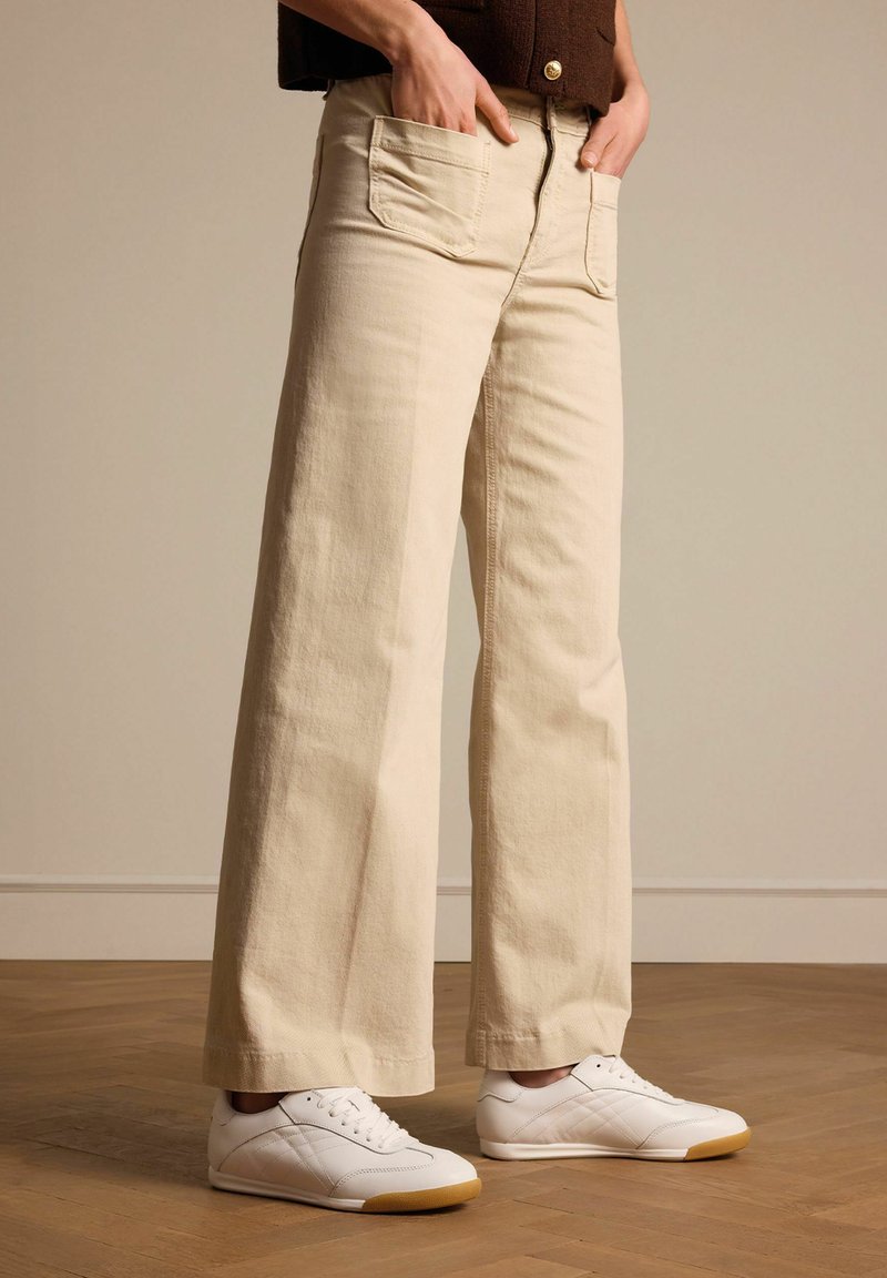 Beige wijde jeans van denim, met voorzakken en een ritsopening, gecombineerd met witte sneakers op een houten vloer.