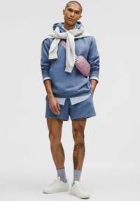 Jasnoniebieska bluza z kapturem, szary sweter związany na szyi, niebieskie szorty, różowa torba na ramię. Białe sneakersy i wzorzyste skarpetki dopełniają wygląd.