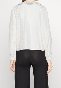 White blouse featuring a floral textured pattern, long sleeves, and a blue-stitched collar. The back is slightly cropped.