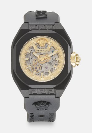 Reloj Versace con correa de silicona negra, caja octagonal, esfera esquelética en tono dorado y acentos con patrón de llave griega. Presenta detalle de cabeza de león.