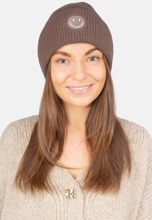 Bonnet marron tricoté avec une texture côtelée et un patch circulaire représentant un visage souriant à l'avant. Porté avec un pull beige clair.