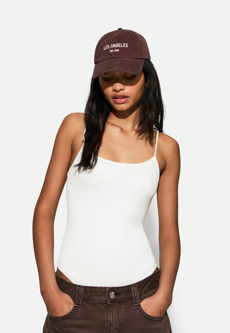 Bijela bodysuit sa tankim naramenicama, tamnosmeđi traper šortsevi i burgundska kapa s izvezenim natpisom "LOS ANGELES EST. 1998" u bijeloj boji.