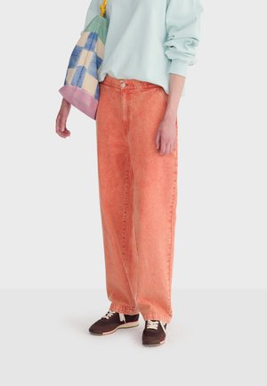 Osoba oblečená v voľných, na vysoký pás strihaných oranžových rifle s efektom vyprania, svetlomodrej mikine s vyhrnutými rukávmi, čiernych teniskách a nesúca viacfarebnú tašku.