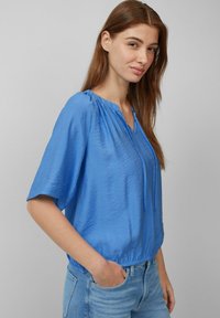 Top azzurro a maniche corte in tessuto leggero e stropicciato, con scollatura a V e dettaglio con cordoncino. Abbinato a jeans di denim blu chiaro.