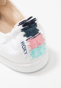 White slip-on sneaker featuring textured fabric, embroidered accents in navy, pink, and teal, with "ROXY" tag on the side. Rubber sole.