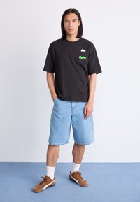 Black cotton t-shirt with a green "Puma" logo and pocket detail, paired with light blue denim shorts and brown sneakers.