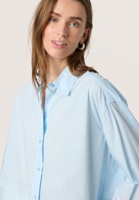 Cămașă oversized de culoare albastră deschisă, cu guler clasic și mâneci largi, fabricată dintr-un material moale din bumbac. Prezintă detalii discrete ale nasturilor.