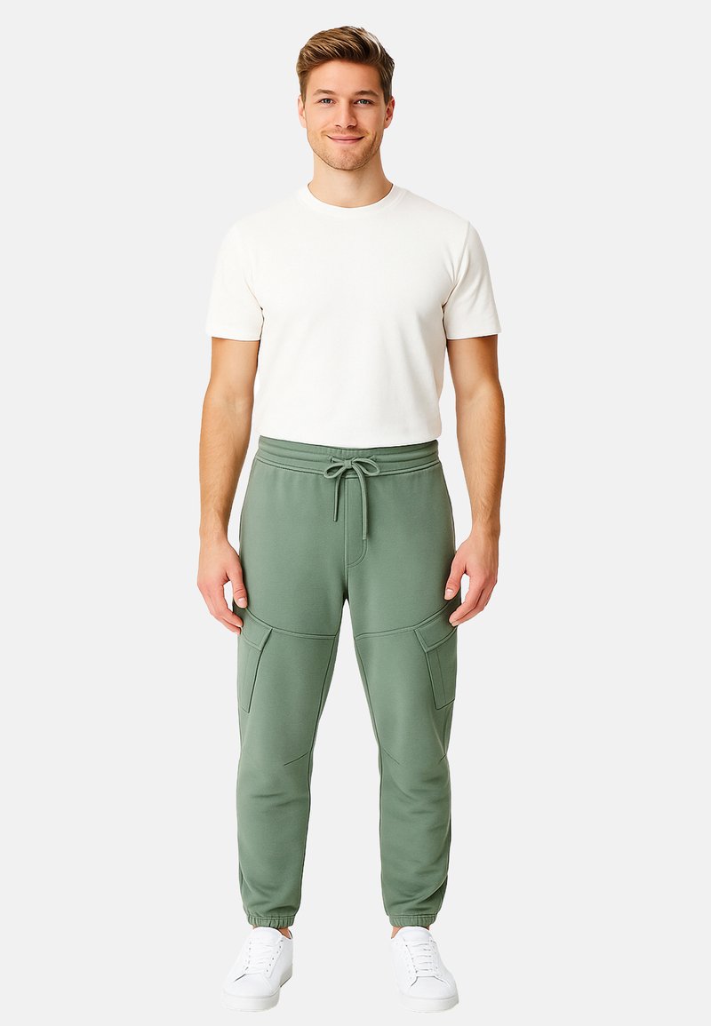 Mann steht da, trägt ein weißes T-Shirt mit kurzen Ärmeln, grüne Cargo-Jogginghosen mit Kordelzug und weiße Sneaker, vor einem einfachen hellen Hintergrund.
