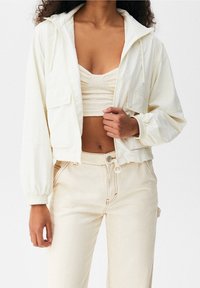 Lekka kremowa krótka kurtka z elastycznymi mankietami, dwoma przednimi kieszeniami oraz kapturem ze sznurkiem, zestawiona z dopasowanym prążkowanym topem i spodniami.