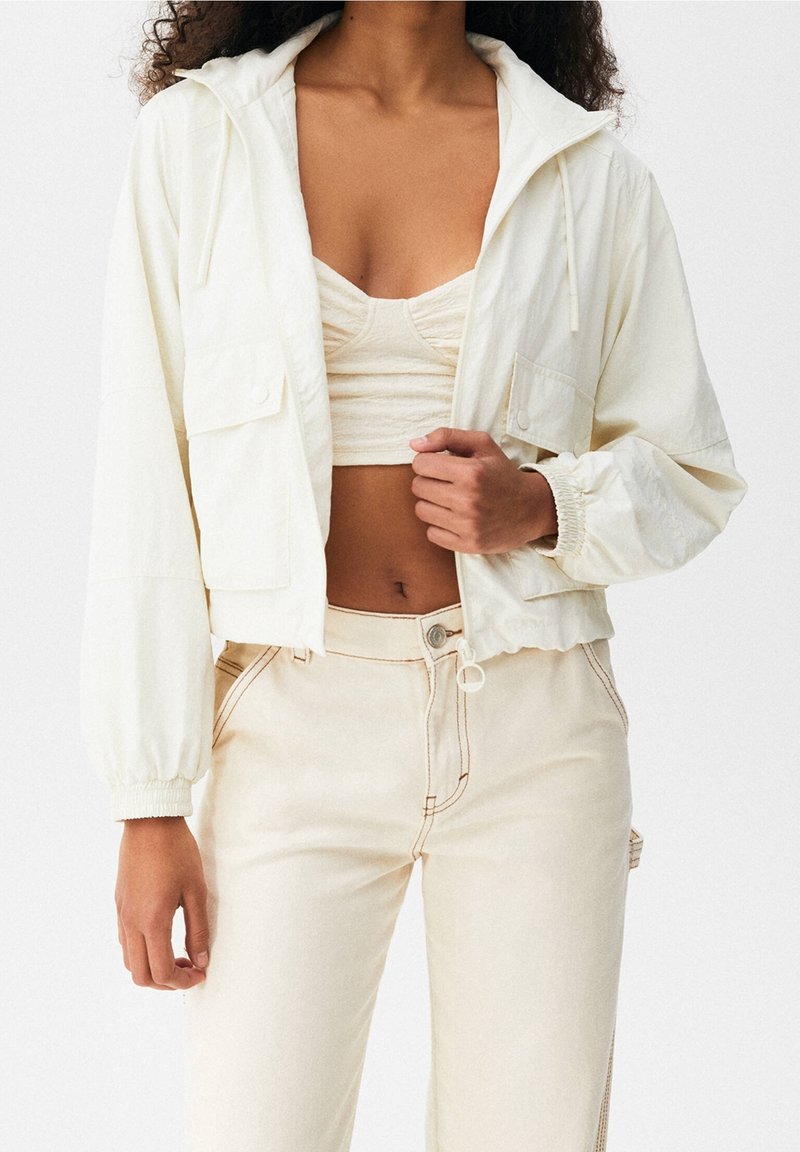 Lekka kremowa krótka kurtka z elastycznymi mankietami, dwoma przednimi kieszeniami oraz kapturem ze sznurkiem, zestawiona z dopasowanym prążkowanym topem i spodniami.