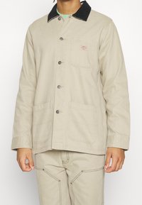 Beige canvas jacket with black corduroy collar, button closure, two front pockets, and contrasting stitching details.