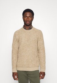 Maglione lavorato a maglia beige con un motivo testurizzato, scollo rotondo e polsini a coste, indossato sopra una camicia a quadri. Pantaloni verdi completano l'outfit.