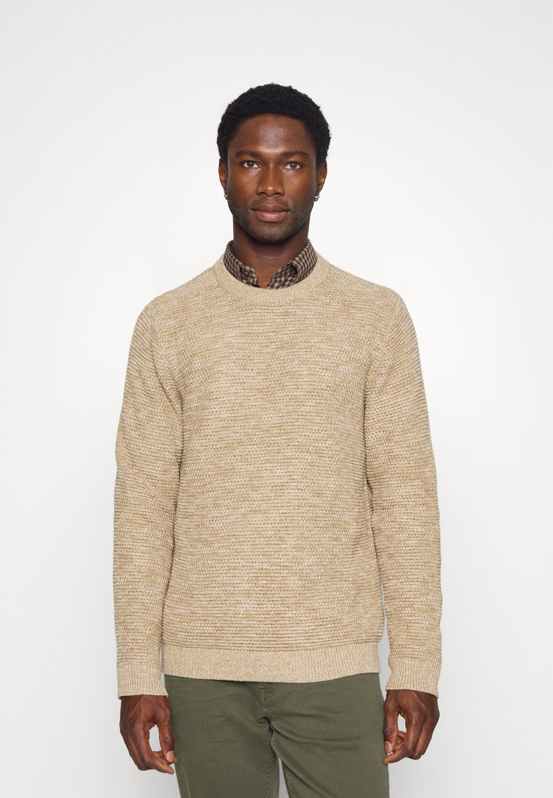 Maglione lavorato a maglia beige con un motivo testurizzato, scollo rotondo e polsini a coste, indossato sopra una camicia a quadri. Pantaloni verdi completano l'outfit.