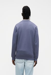 Lilla pusa, millel on soonikust ümar kaelus, pehme kangas, pikad varrukad ja lõdvestunud lõige, siniste teksadega, vaadatuna tagantpoolt.