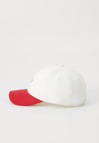 White cap with a red brim, featuring a stitched logo on the front. Made of cotton, it has six panels and ventilation eyelets.