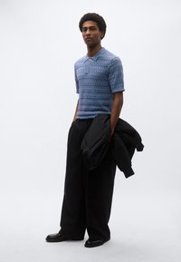 Μπλε κοντομάνικη πόλο μπλούζα με υφή, σε συνδυασμό με άνετο μαύρο παντελόνι και μαύρο σακάκι που κρέμεται από το χέρι.