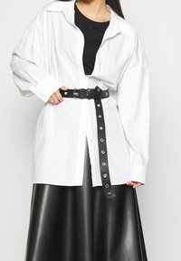 Weißes Oversized-Hemd, darunter ein schwarzes Top, mit einem schwarzen Gürtel mit metallischen Details tailliert, kombiniert mit einem schwarzen Lederrock.