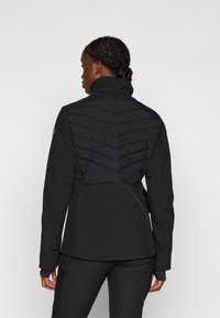 Veste isolante noire avec un col haut, un motif en chevrons matelassé à l'arrière, et un design cintré avec des manches longues.