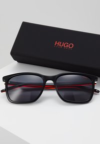Ochelari de soare negri cu lentile gri, design cu ramă pătrată, accente roșii pe tâmple și un etui elegant negru, etichetat "HUGO."
