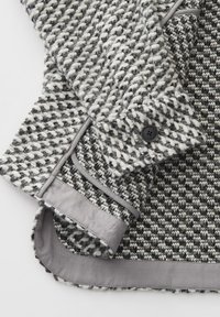 Gewebter Mantel in Schwarz und Weiß mit strukturiertem Oberflächen, ausgestattet mit einem grauen Satinfutter und Knopfakzenten. Strukturierter Kragen und Manschetten sind sichtbar.