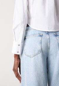 Camicia bianca a bottone con design corto, caratterizzata da maniche lunghe e bottoni a pressione ai polsini; abbinata a jeans a vita alta di un blu chiaro.